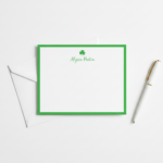 Flatlay of a green‑border shamrock stationery note card with a white envelope and a white pen with a gold tip on a white background, styled for St. Patrick’s Day notes.