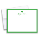 Masculine desk flatlay with a green‑border shamrock note card reading ‘TIM MULLANEY,’ a white envelope, white pen with gold tip, leather watch, black eyeglasses, and a white mug of black coffee on a dark wood surface.