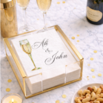 Elegant bar flatlay showcasing personalized champagne cocktail napkins alongside champagne flutes, gold barware, and soft candlelight for a celebratory wedding or engagement party setting.