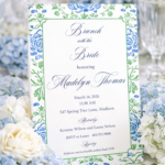 Bridal shower invitation with blue and green floral border displayed on table with blue and white hydrangeas and elegant table setting