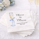 Personalized bridal shower napkins with blue ribbon champagne design styled on white table with champagne flutes and florals
